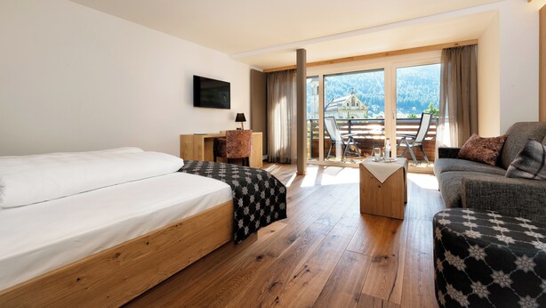 Habitación de hotel con una cama grande, un escritorio, una silla y un sofá. Tiene un balcón con vistas a las montañas.