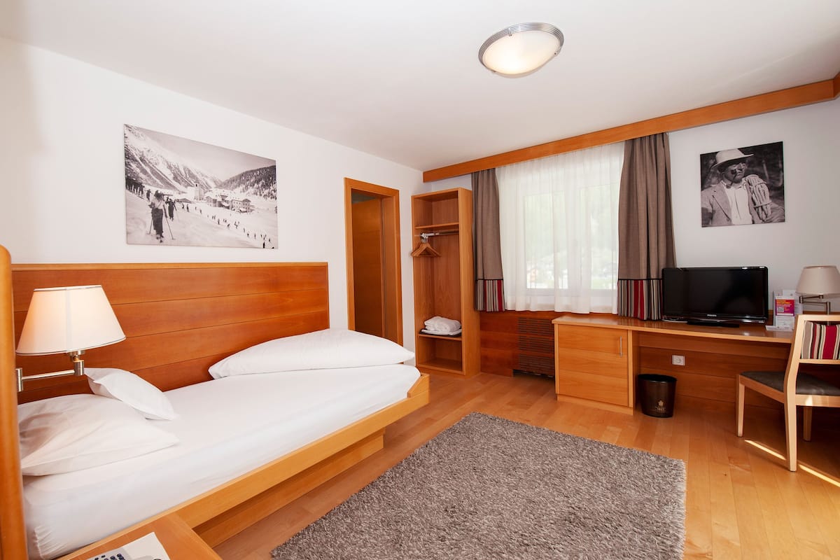 A hotel room with a wooden bed, a desk with a television, a coat rack, and a window with curtains.