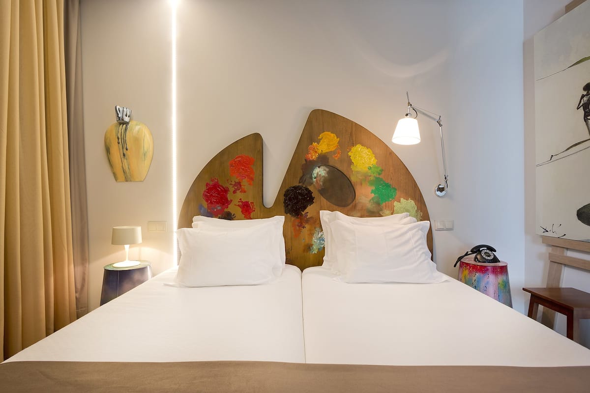 Ein Hotelzimmer mit Bett, zwei Kissen, einer Nachttischlampe, einem Nachttisch mit Lampe, einem Telefon und einer Wand mit einem Gemälde.