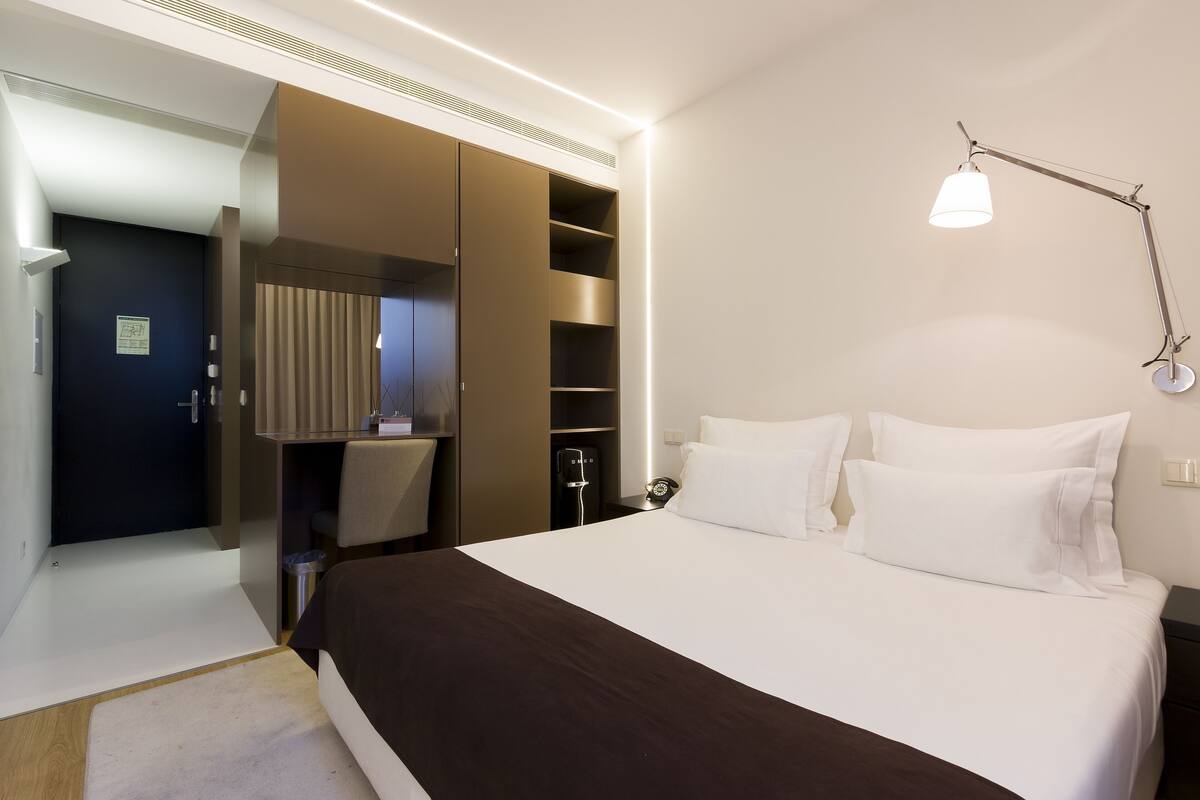 A hotel room with a bed, bedside table, lamp, wardrobe, and a desk with chair.