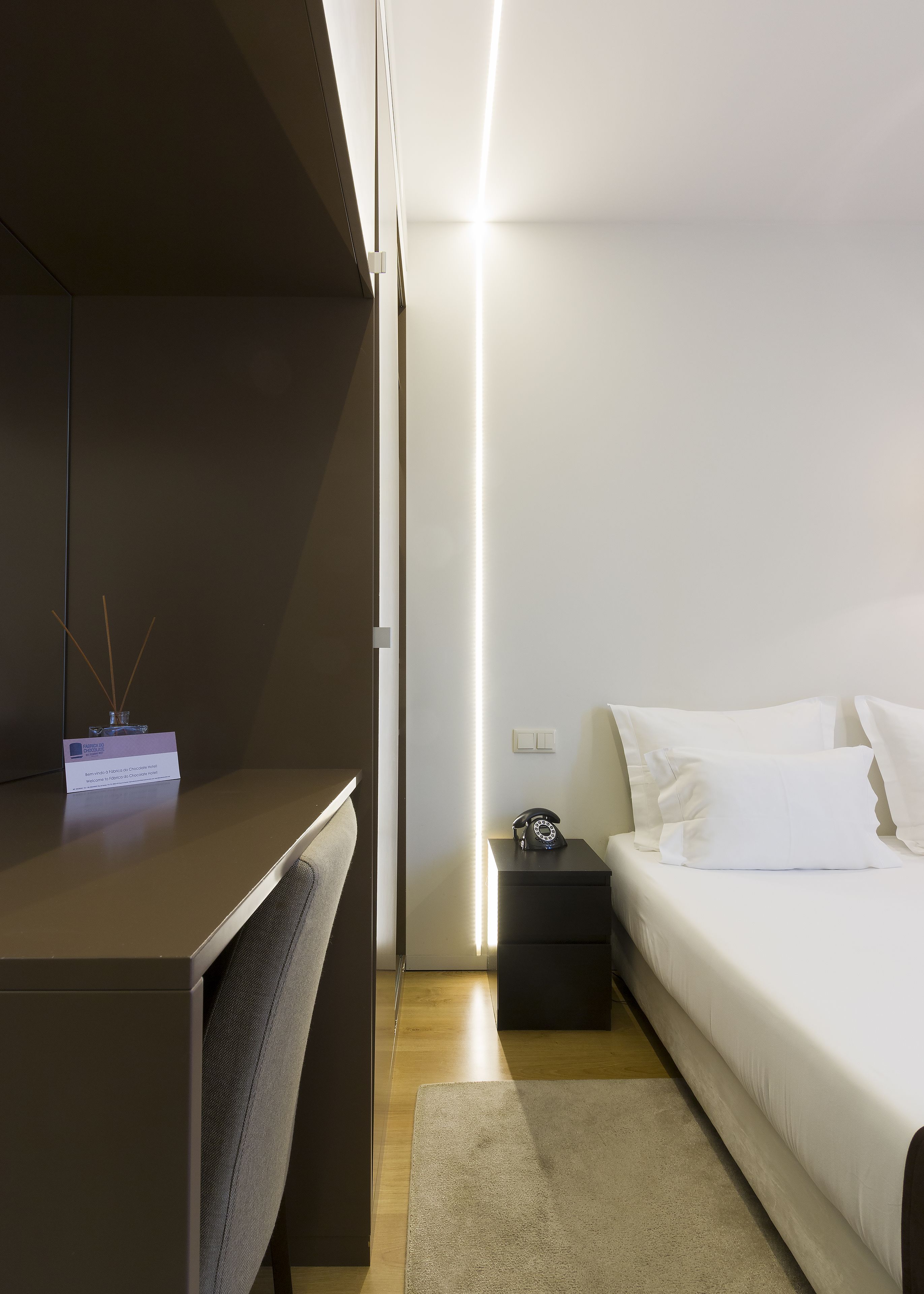 A modern hotel room with a bed, bedside table, and a desk with a lamp.