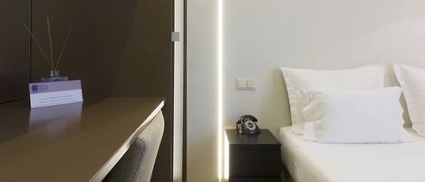 A modern hotel room with a bed, bedside table, and a desk with a lamp.