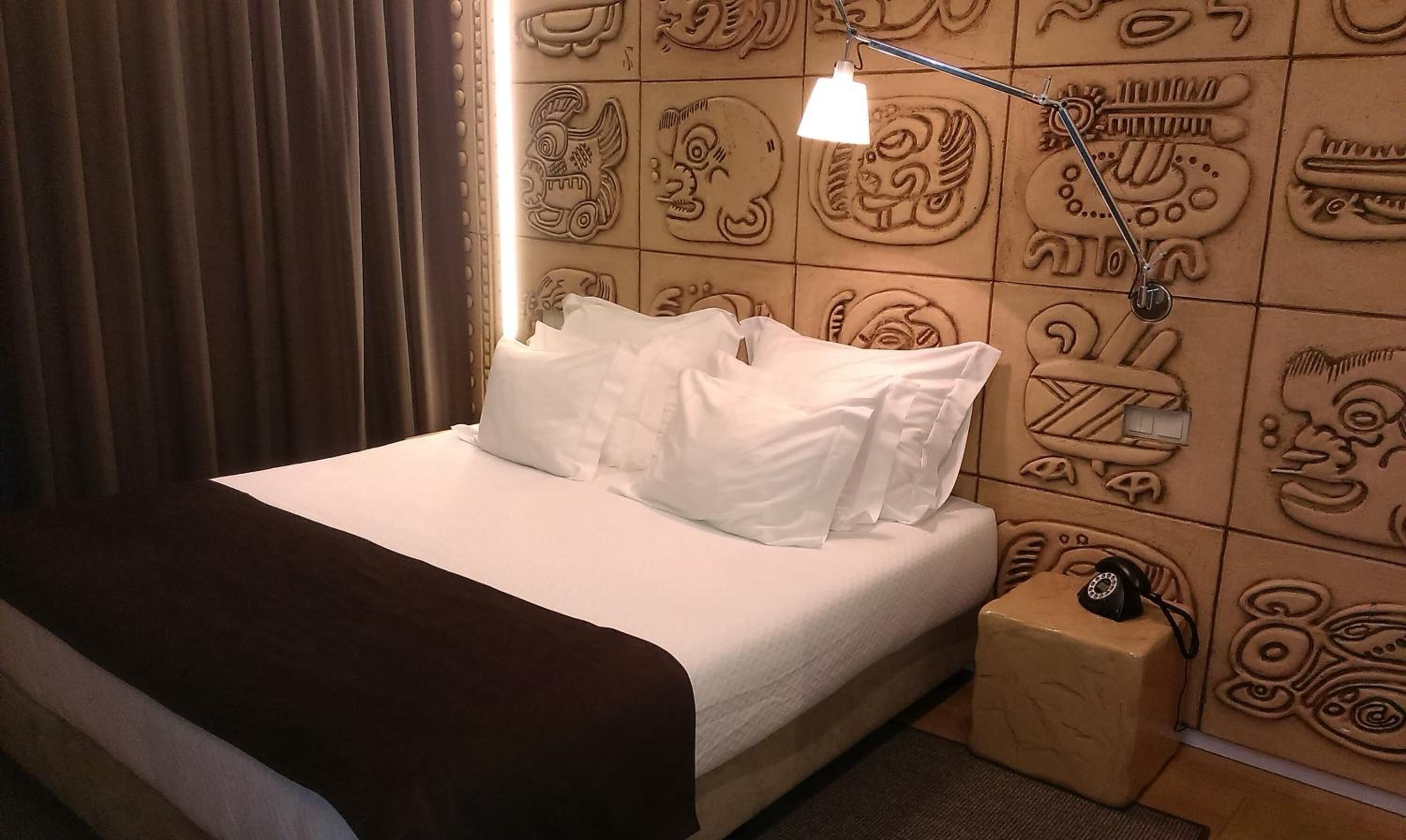 Une chambre d’hôtel comprenant un lit, des oreillers, une table de chevet avec une lampe et un mur orné de motifs sculptés.