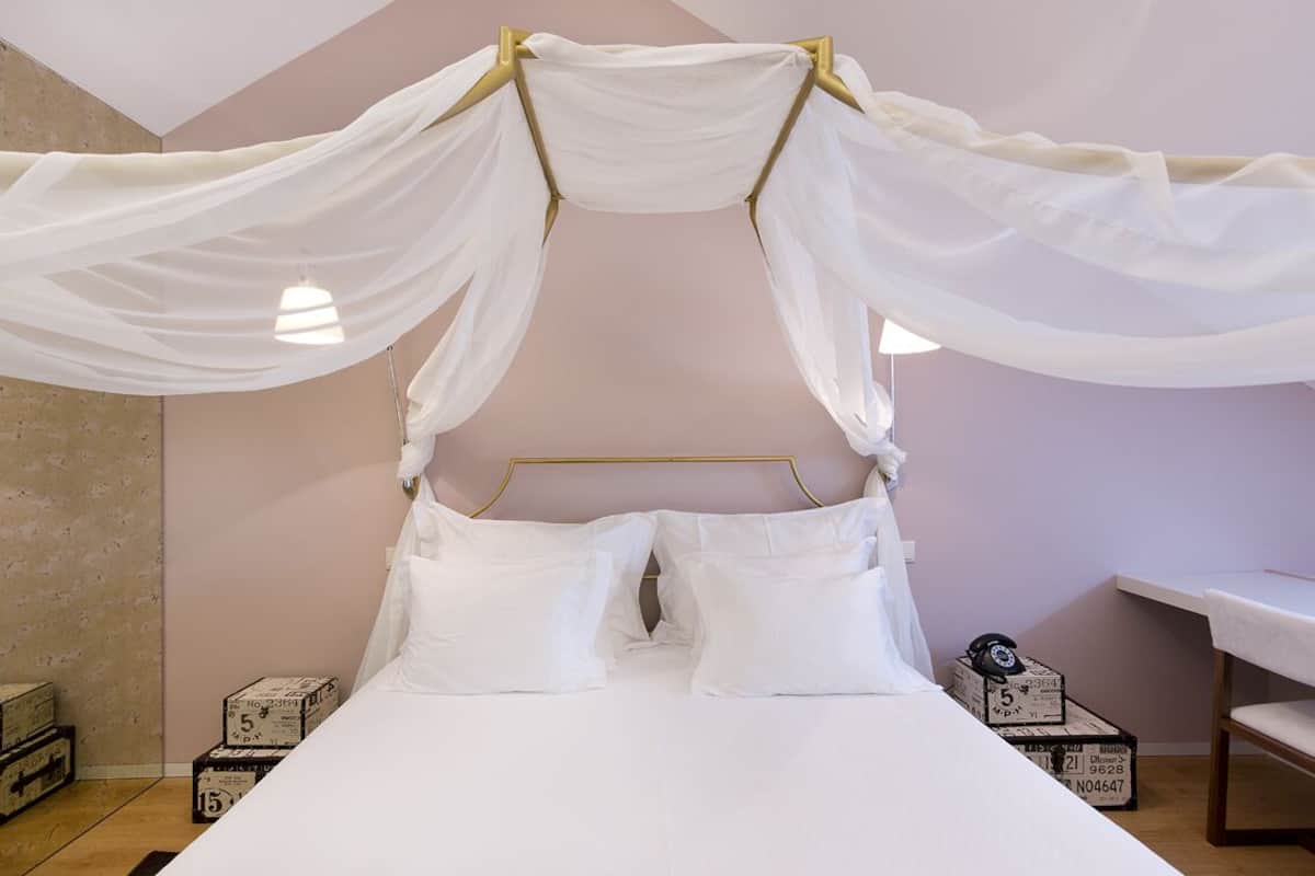 Ein Bett mit Baldachin, weißer Bettwäsche und einem goldenen Kopfteil. Es gibt zwei Nachttische und eine Wandlampe.