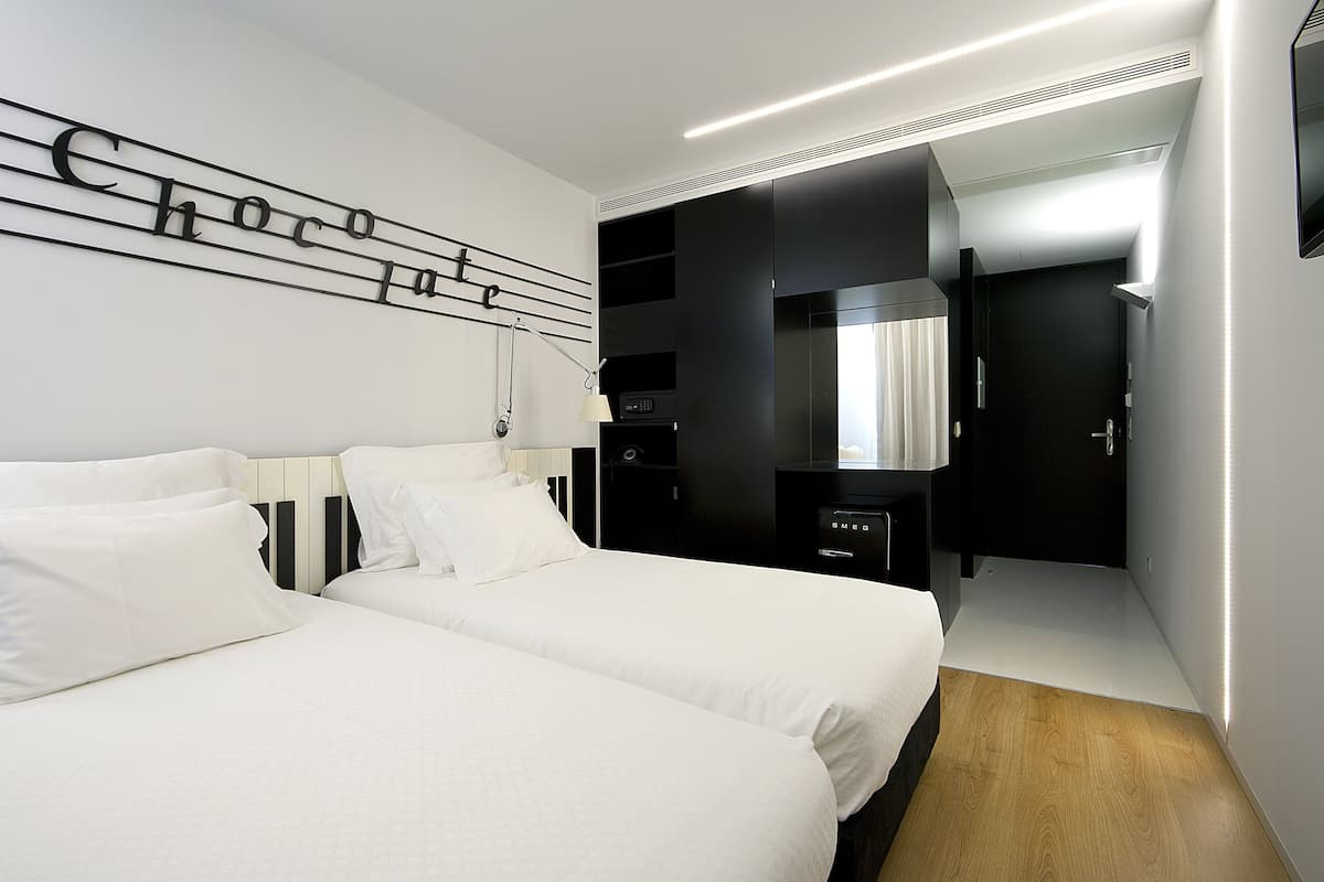 A modern hotel room with two beds, a black wardrobe, a mirror, and a wall-mounted light fixture.
