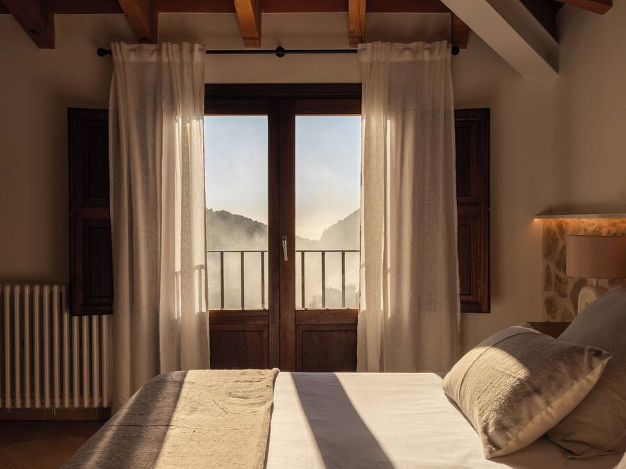 A bedroom with a bed, curtains, a view of mountains, a radiator, and a stone wall.