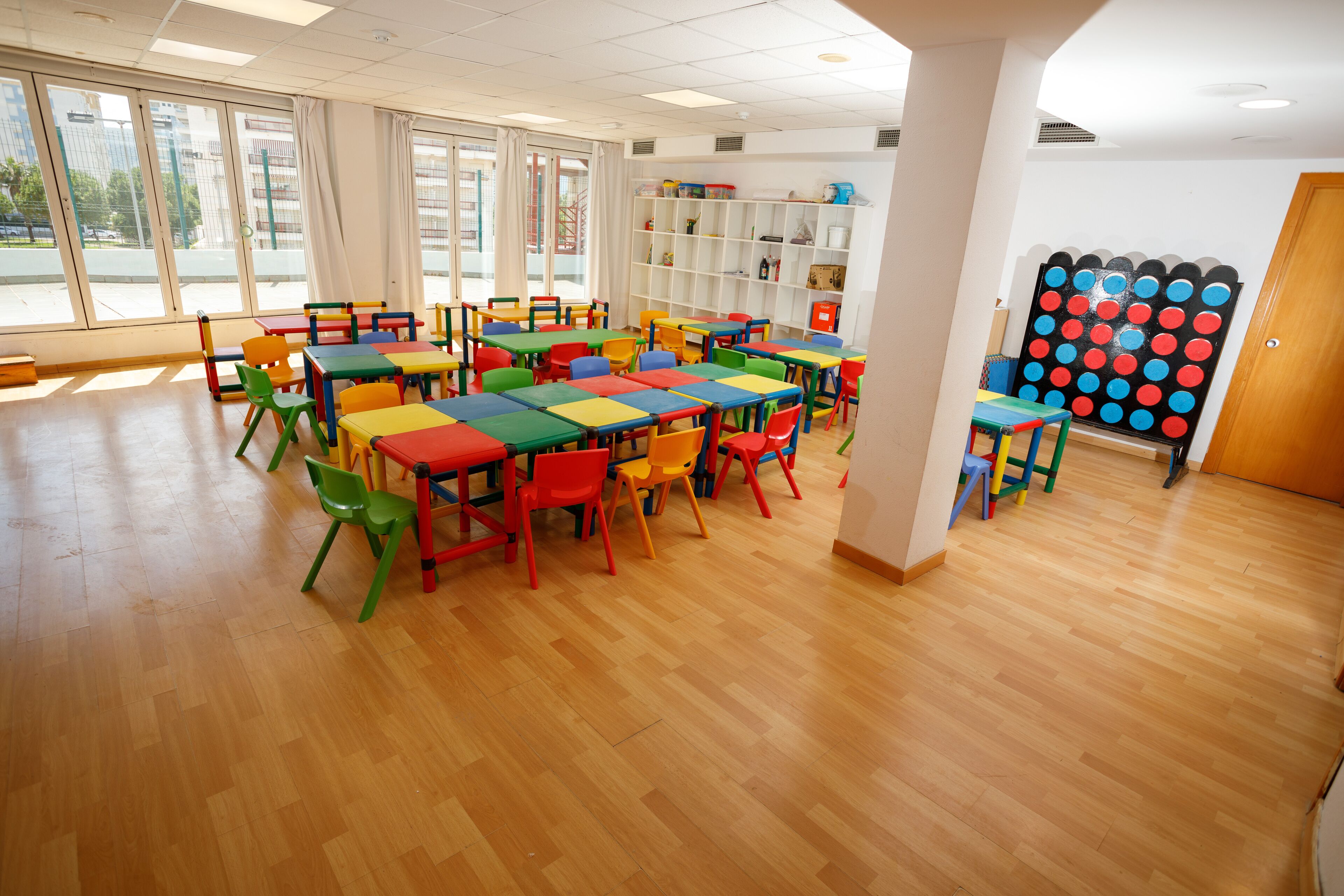 Un aula con mesas y sillas coloridas, ventanas amplias y una estantería.