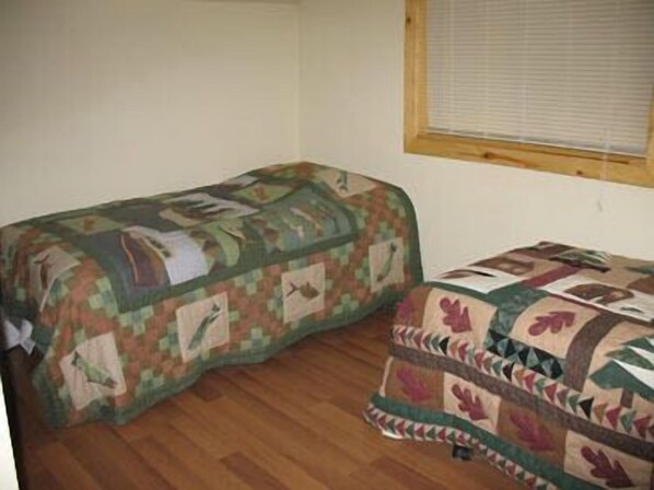 A room with two beds covered in patterned quilts, wooden flooring, and a window with blinds.