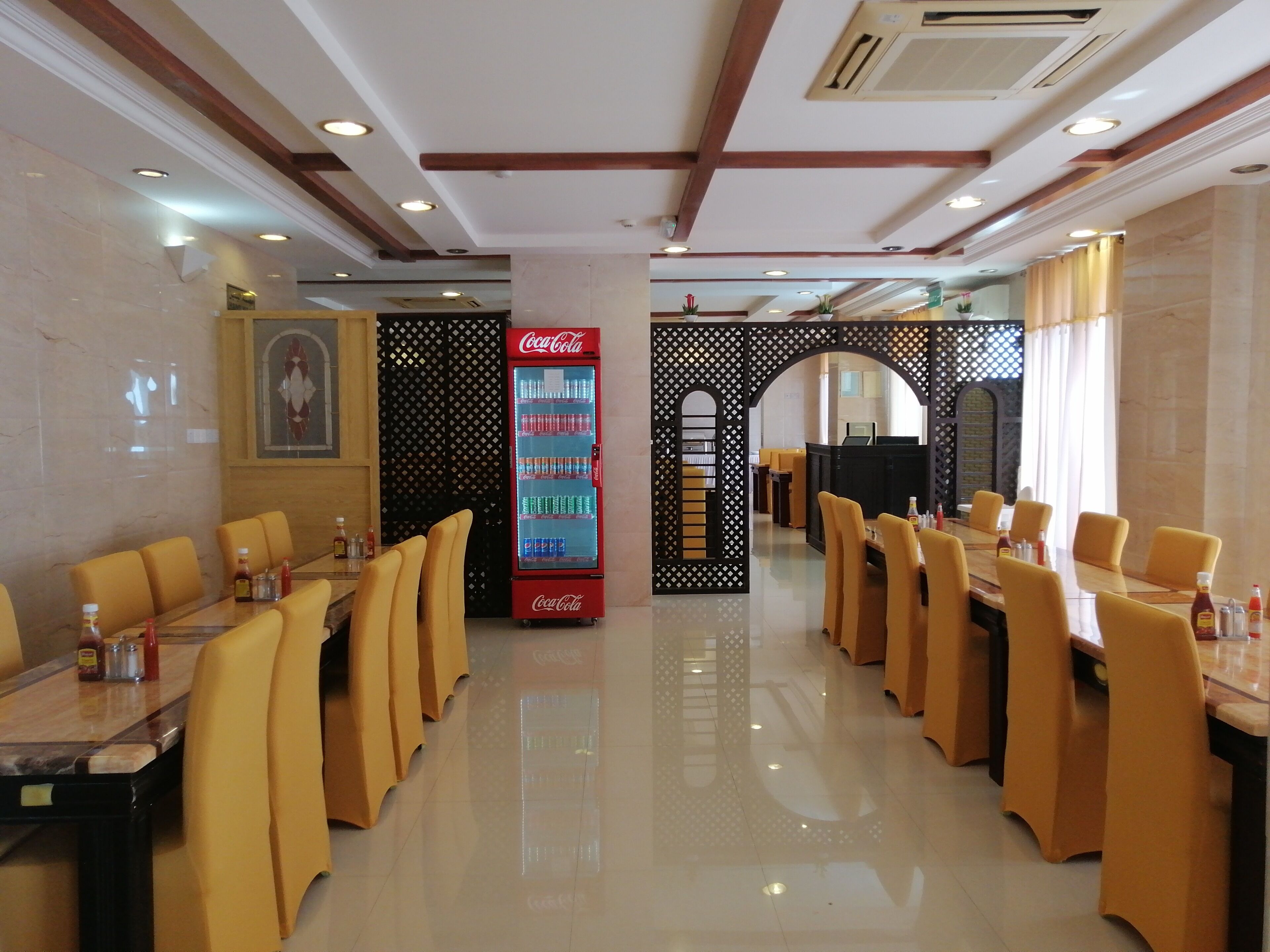 A restaurant with multiple dining tables and chairs, a Coca-Cola refrigerator, and a decorative wall with an archway.