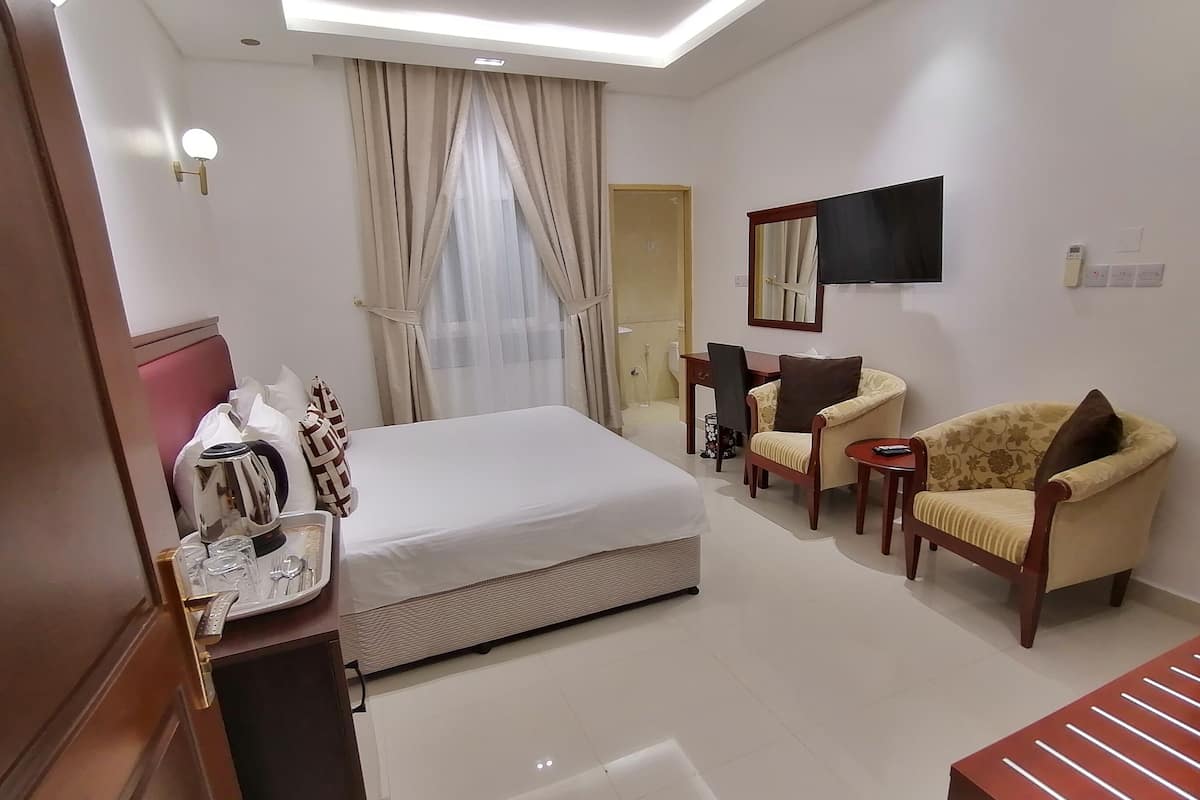 A hotel room with a bed, a desk, a chair, a TV, and a bedside table with a kettle and glasses.
