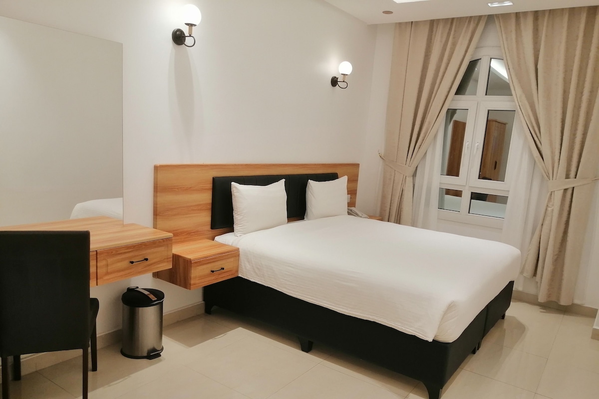 Een hotelkamer met een houten hoofdeinde, een bed met wit beddengoed, een bureau met een stoel en een prullenbak.