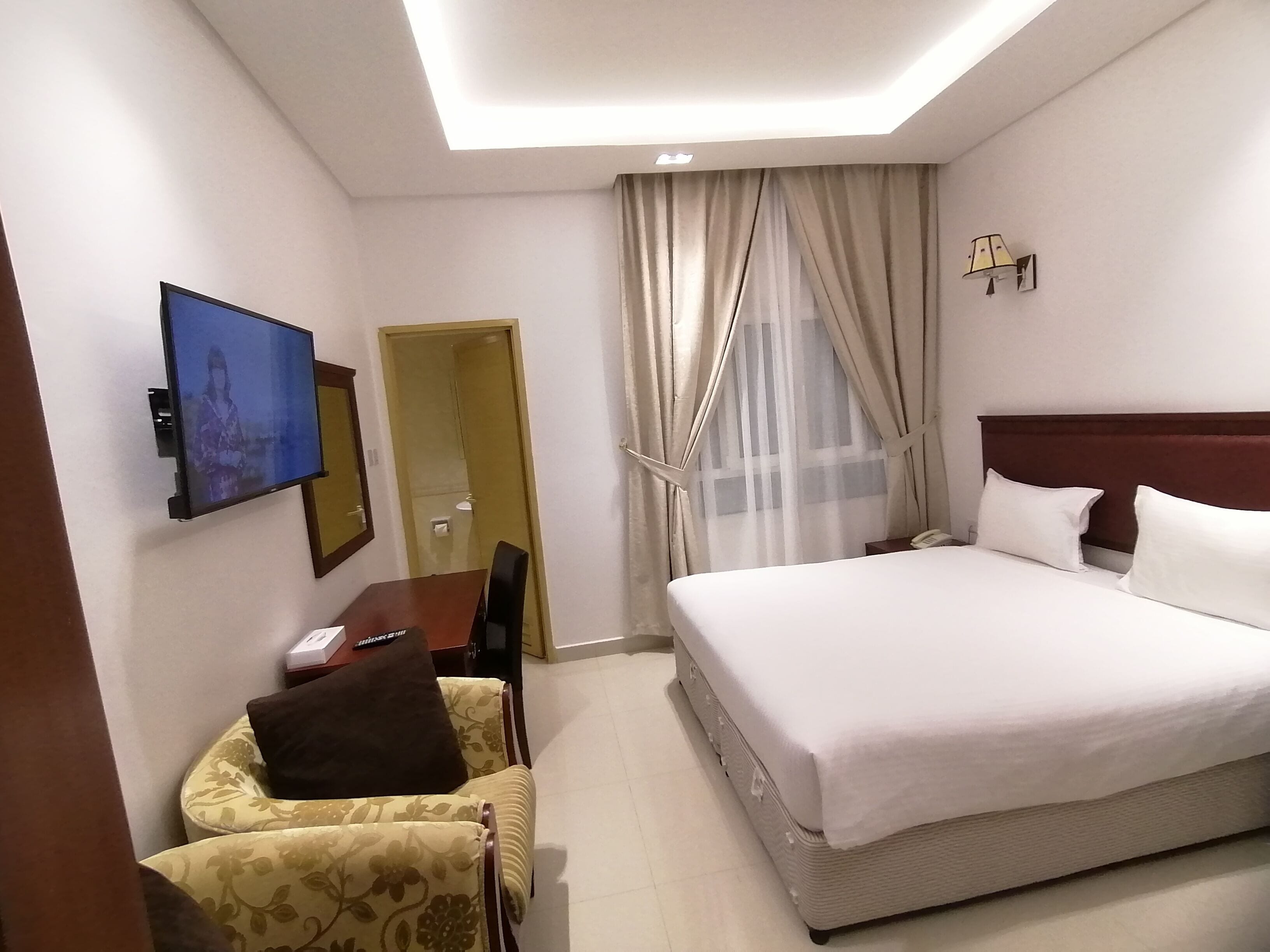 A hotel room with a bed, a TV mounted on the wall, a desk with a chair, and a window with curtains.
