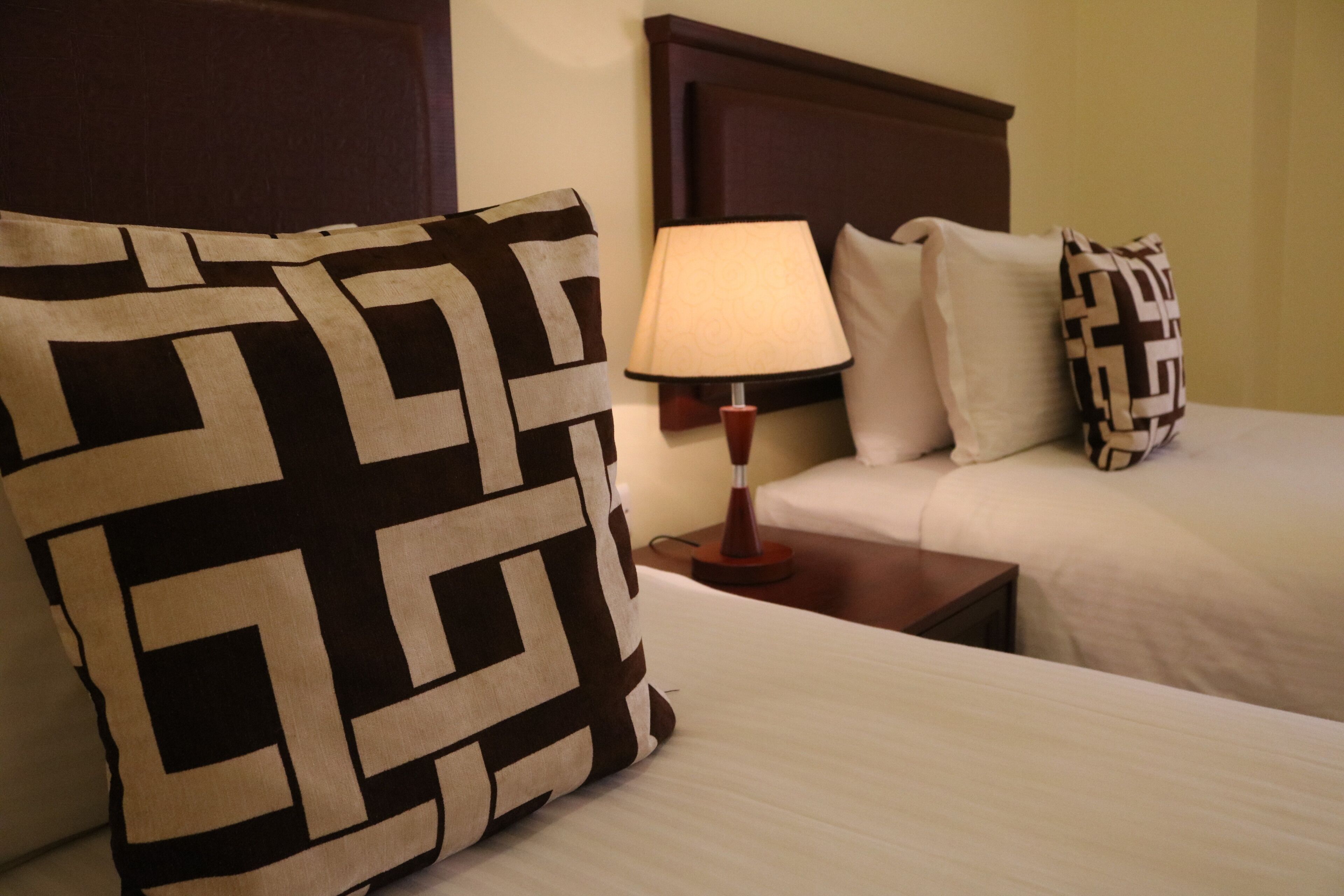 A hotel room with two beds, a nightstand with a lamp, and patterned pillows.
