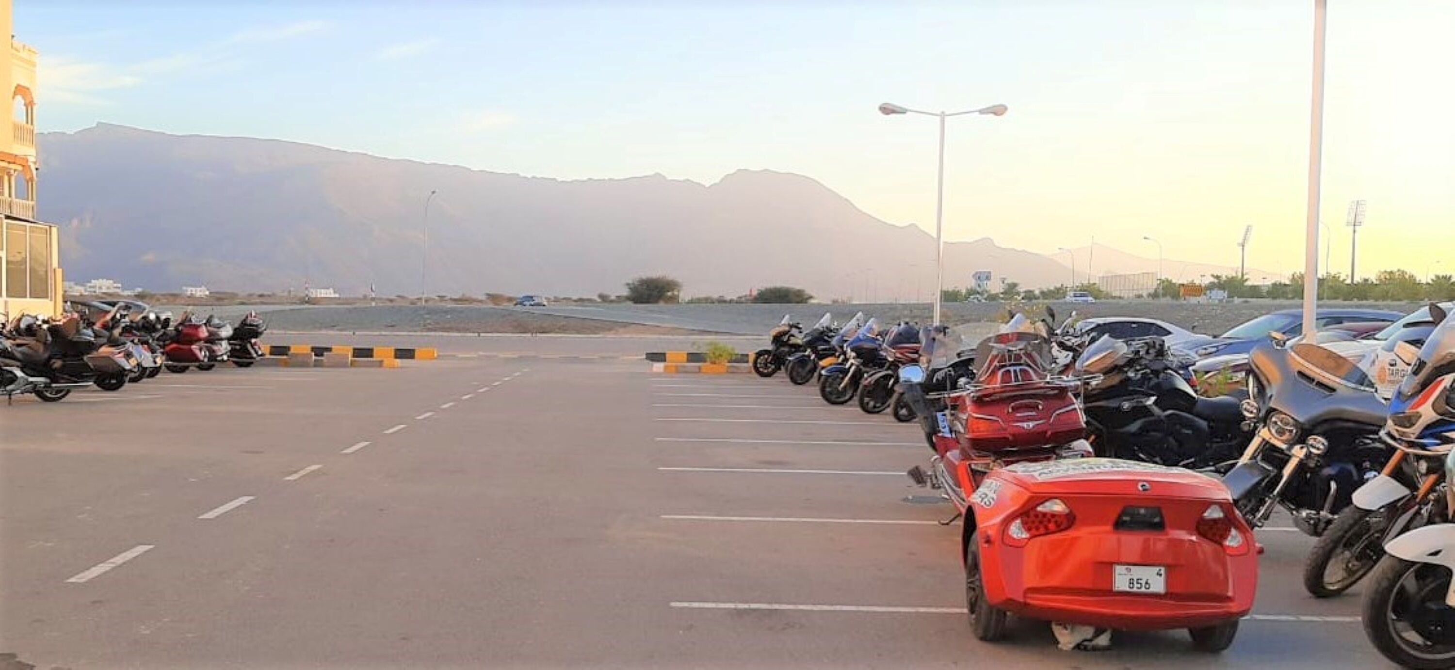 Un estacionamiento con motocicletas y un auto rojo, con montañas al fondo.