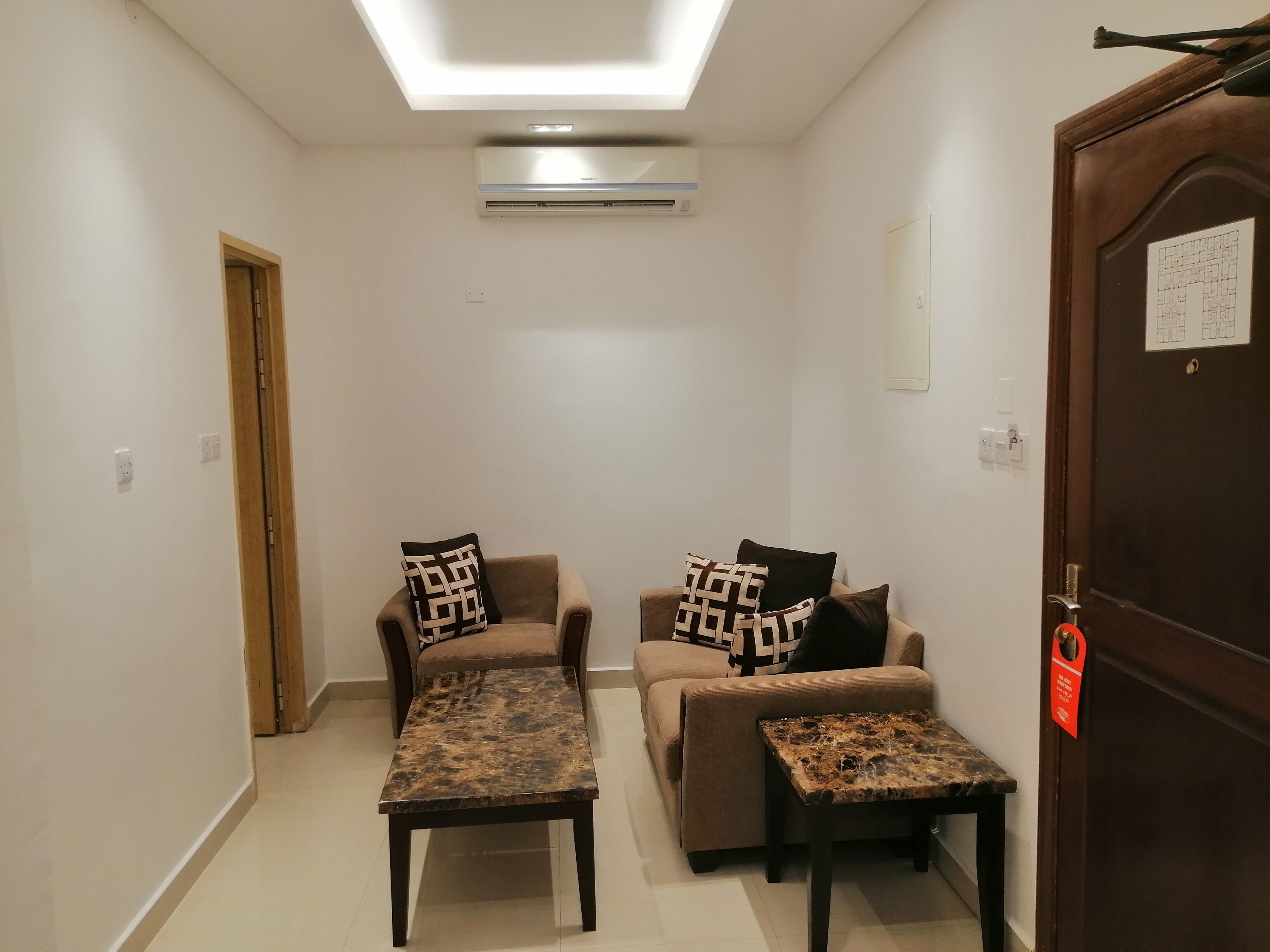 A modern living room with a sofa, two armchairs, a coffee table, and a small side table. There is a ceiling light, an air conditioning unit, and a door with a lock.