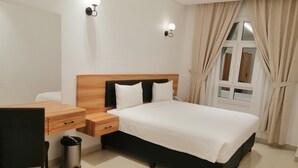 Habitació d'hotel amb un capçal de llit de fusta, un llit amb roba de llit blanca, una taula de treball amb una cadira i un cubell de brossa.