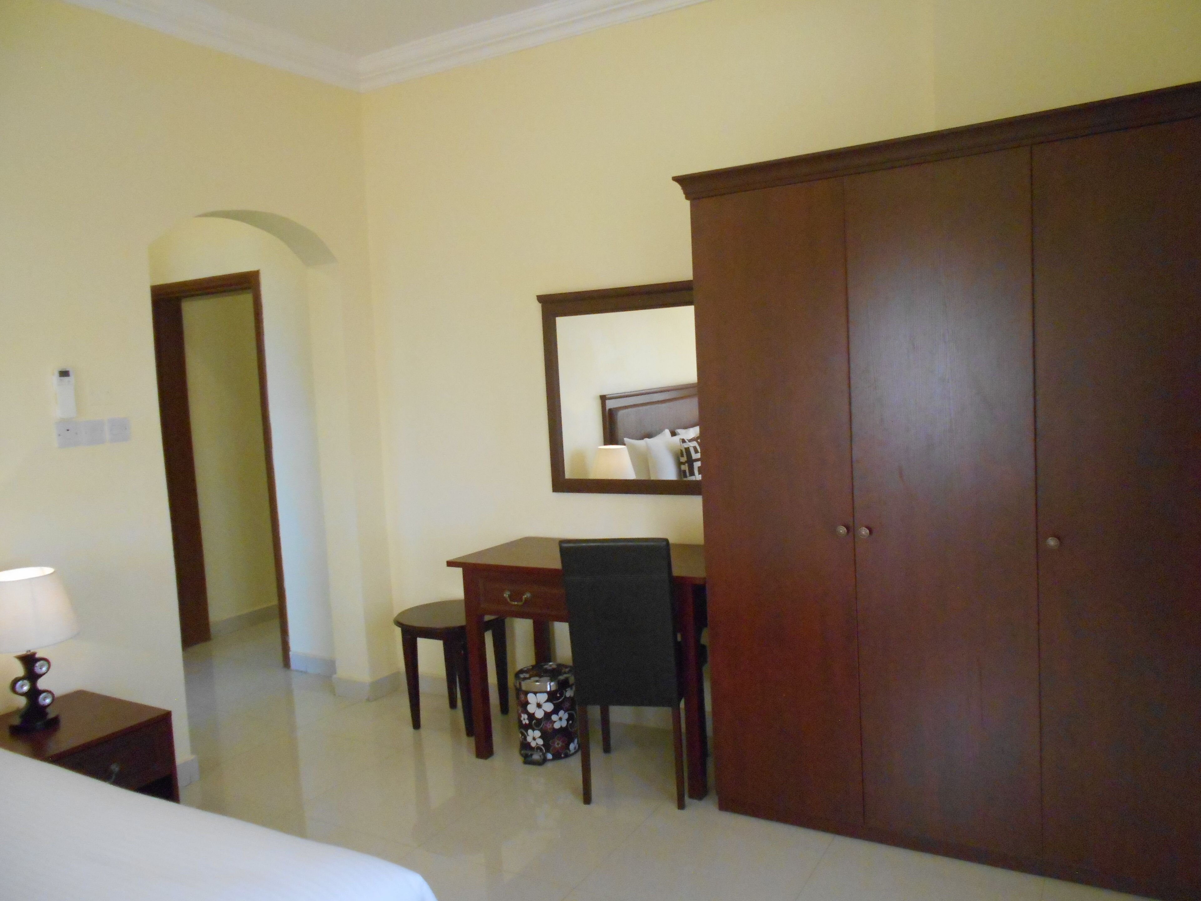A hotel room with a bed, a desk, a chair, a wardrobe, and a small table with a lamp.