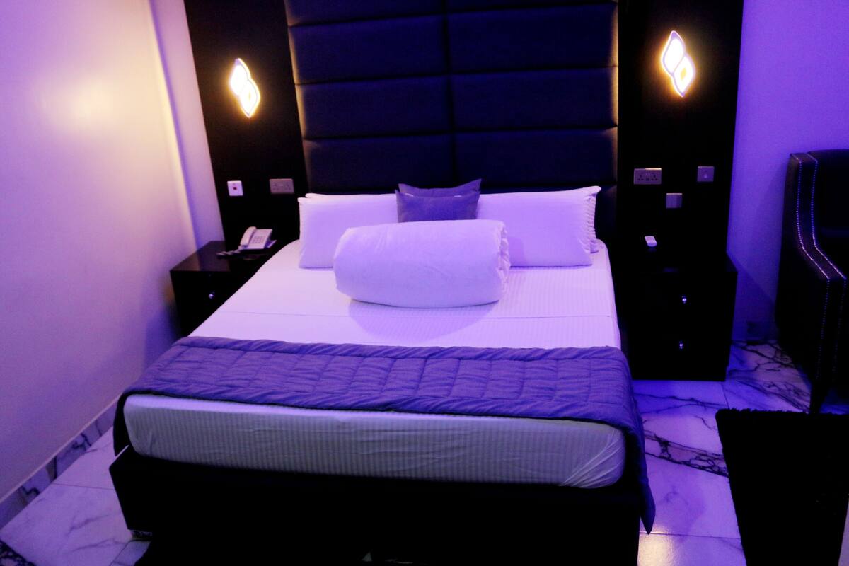 A bed with a headboard, a bedside table with a phone, and a nightstand with a lamp.