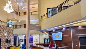 A hotel reception area with a wooden reception desk, a large screen displaying the hotel name, and a staircase leading to an upper level. - Parkview Astoria Hotel (Lagos)