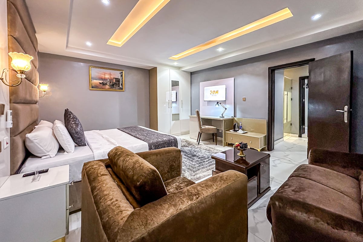 A modern hotel room with a bed, a sofa, a desk with a chair, a TV, and a wardrobe.