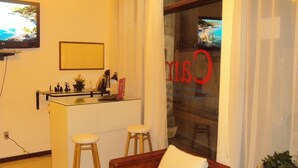 A compact living space with a kitchenette, a sofa with cushions, a small stool, and a television mounted on the wall. - Pousada Camburi (Morro de Sao Paulo)