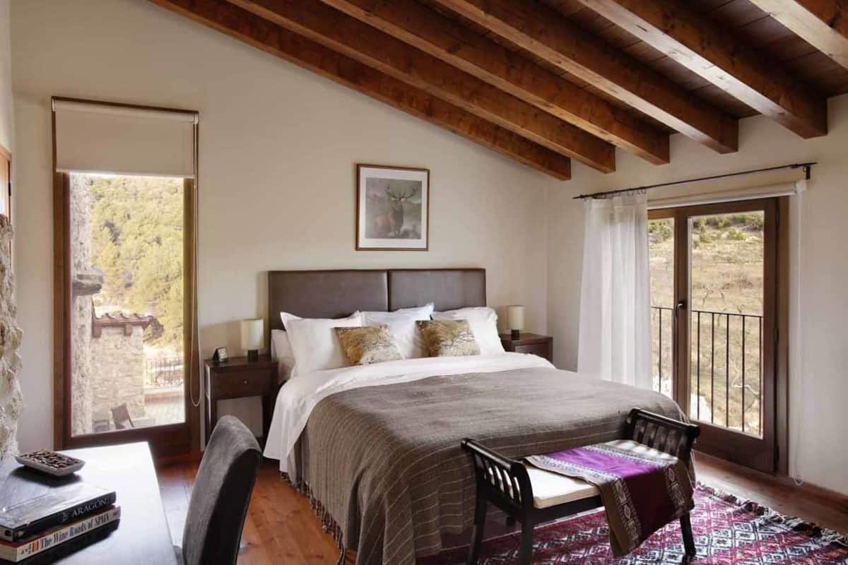 A bedroom with a large bed, wooden beams, a window with a view, and a desk with books.