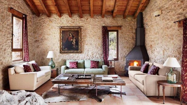 Un salón rústico con paredes de piedra, vigas de madera, chimenea y dos sofás con cojines.