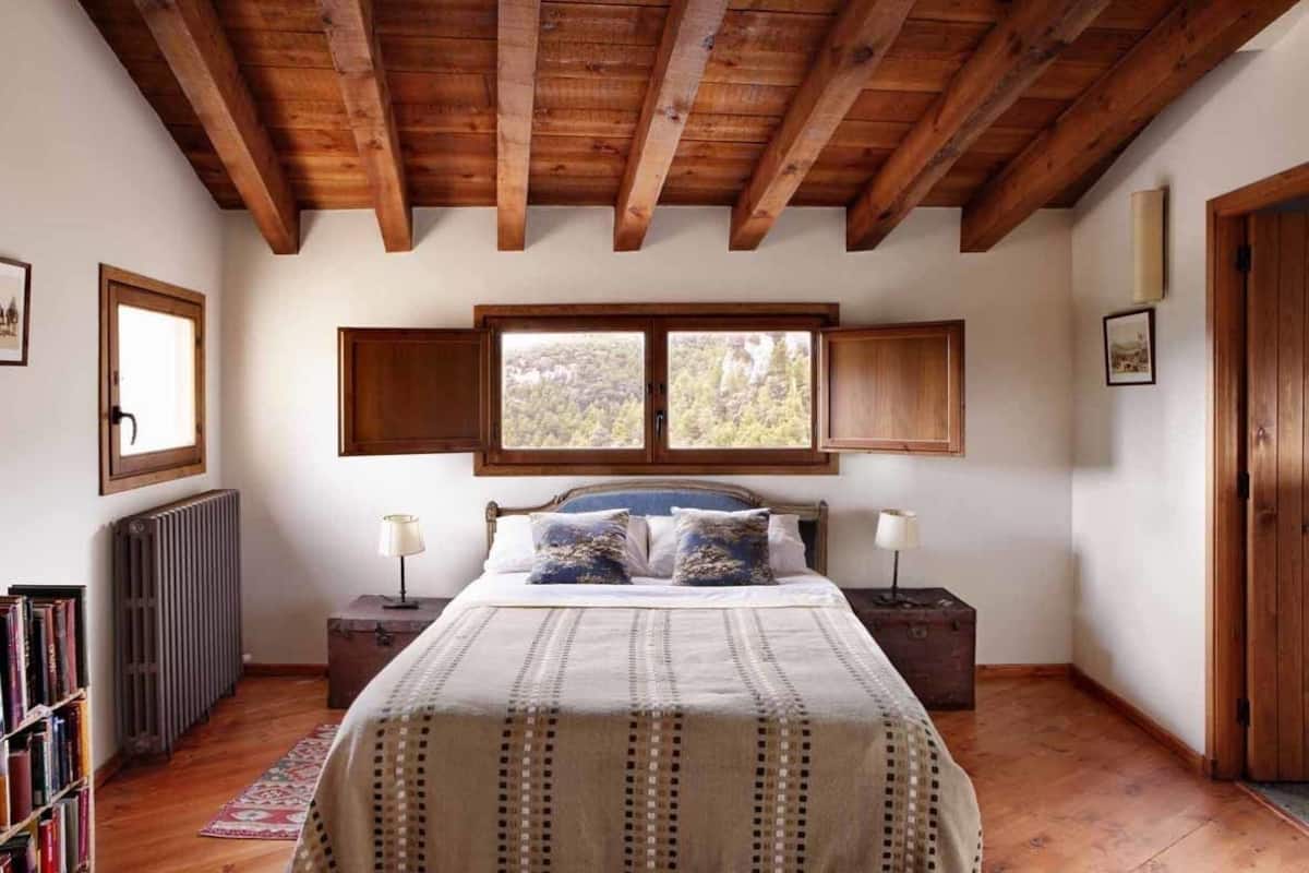 A bedroom with a wooden ceiling, a large bed with a patterned bedspread, two bedside tables with lamps, a radiator, and a bookshelf.