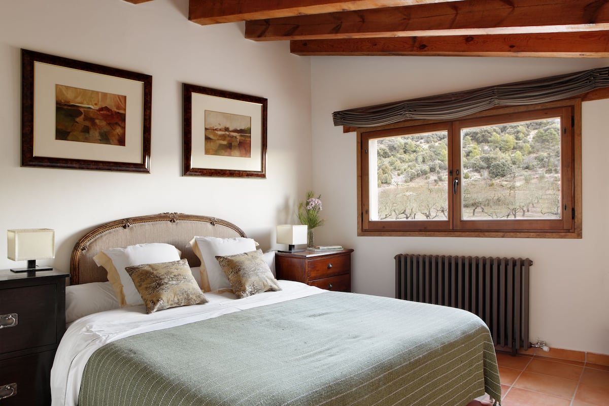 A bedroom with a bed, two framed pictures on the wall, a nightstand with a lamp, and a window with a view of trees.