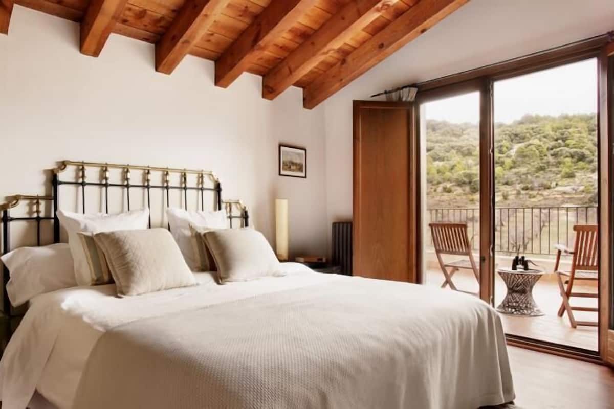 A bedroom with a large bed, wooden ceiling beams, a balcony with chairs, and a view of a mountainous landscape.