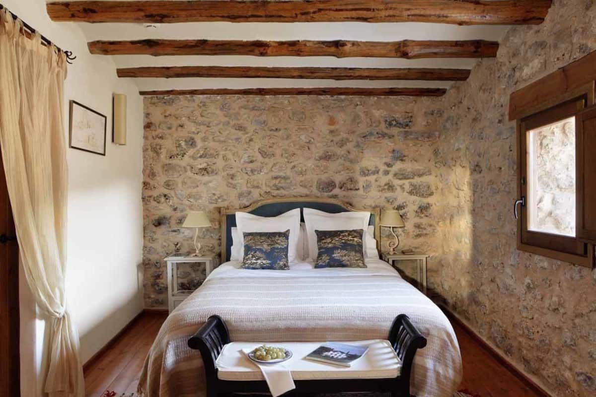 A bedroom with a stone wall, wooden beams, a bed with white bedding, two bedside tables with lamps, and a window.