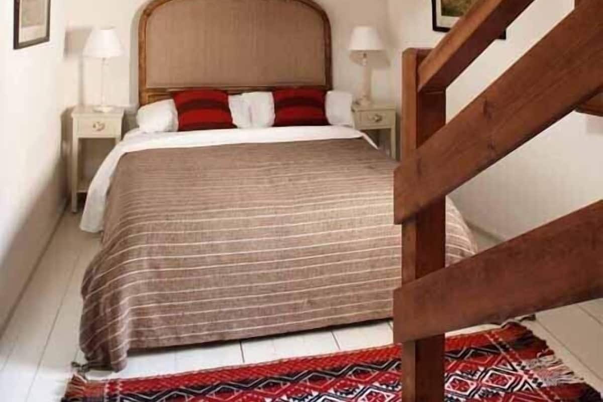 A bedroom with a sloped ceiling, a wooden staircase, a bed with a headboard, bedside tables, and a hanging light fixture.