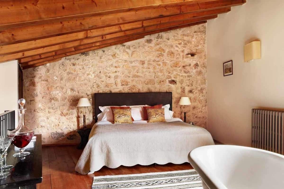 A bedroom with a stone wall, a wooden ceiling, a bed with white bedding, two bedside lamps, and a bathtub.