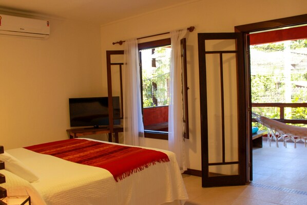 A hotel room with a bed, a television, a balcony with a hammock, and a view of greenery outside. - Via das Pedras Pousada (Cairu)