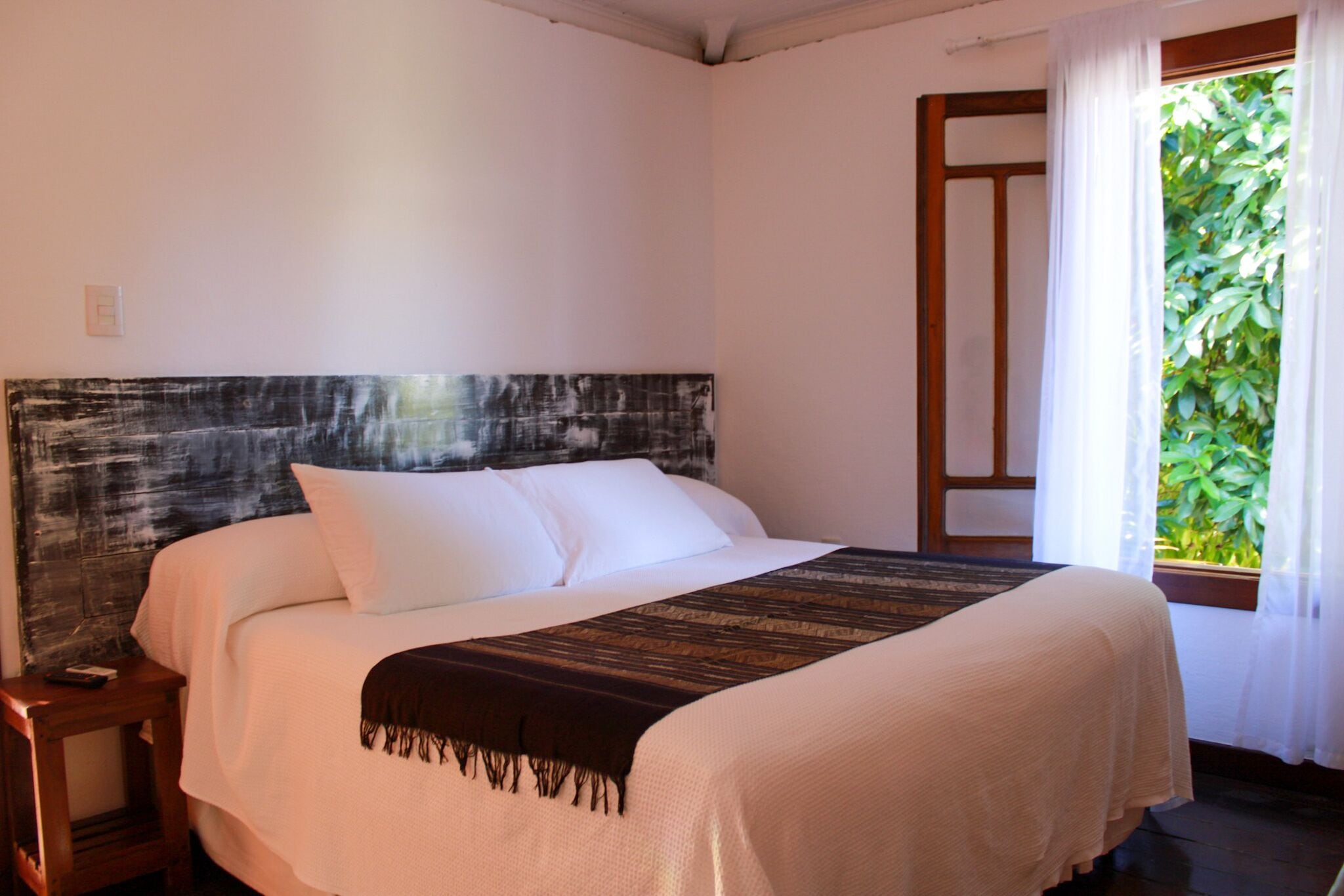 A neatly made bed with white linens and a dark headboard, a wooden nightstand, and a window with sheer curtains and a view of greenery.