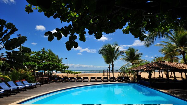 Zona de piscina con sillas de descanso, palmeras y vistas a la playa.
