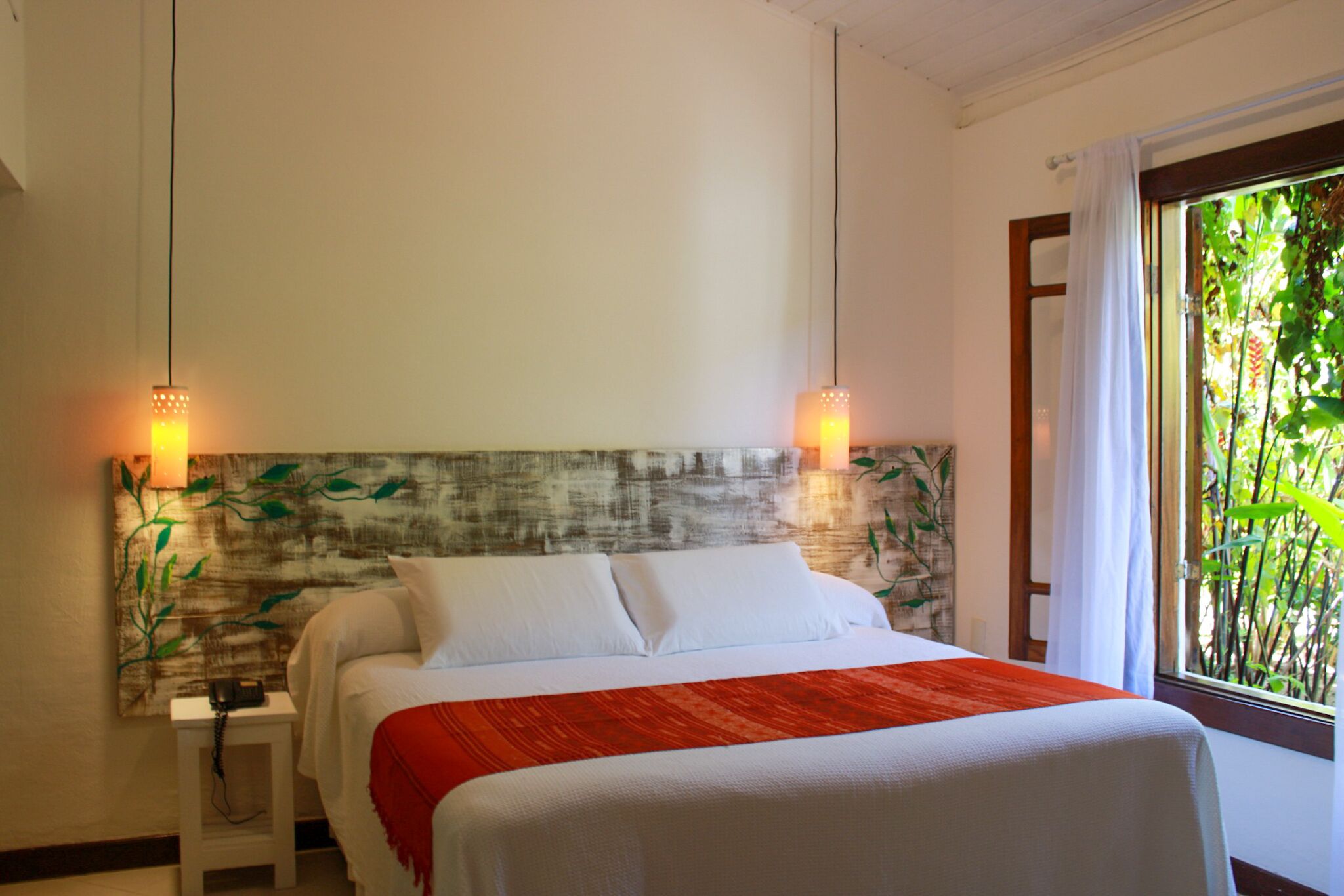 A neatly made bed with white linens and a red accent throw, a wall-mounted lamp, and a window with a view of greenery.