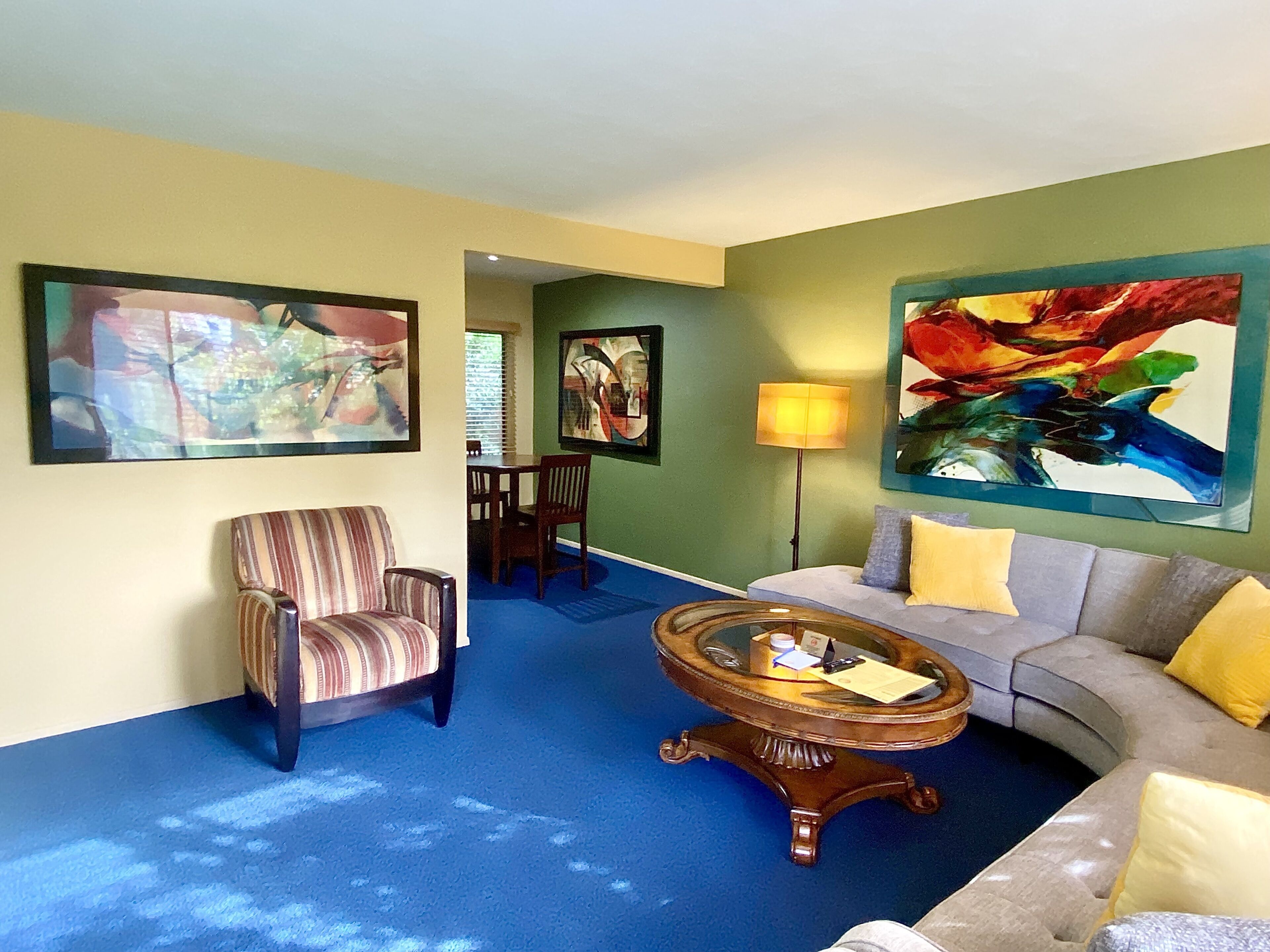 A living room with a blue carpet, a striped armchair, a grey sofa with yellow cushions, a round coffee table, and two large abstract paintings on the walls.