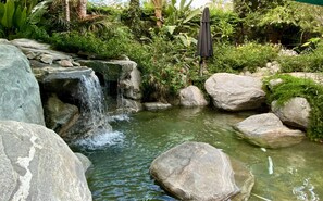 Un jardín con cascada pequeña, rocas grandes y abundante follaje.