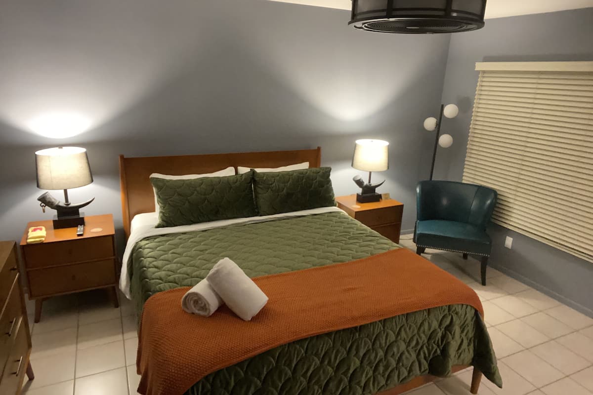 A hotel room with a bed, two bedside lamps, a chair, a window with blinds, and a ceiling light fixture.