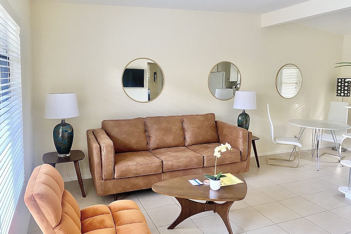 A living room with a brown sofa, two side tables with lamps, a small round table with a plant, and a white dining table with chairs.