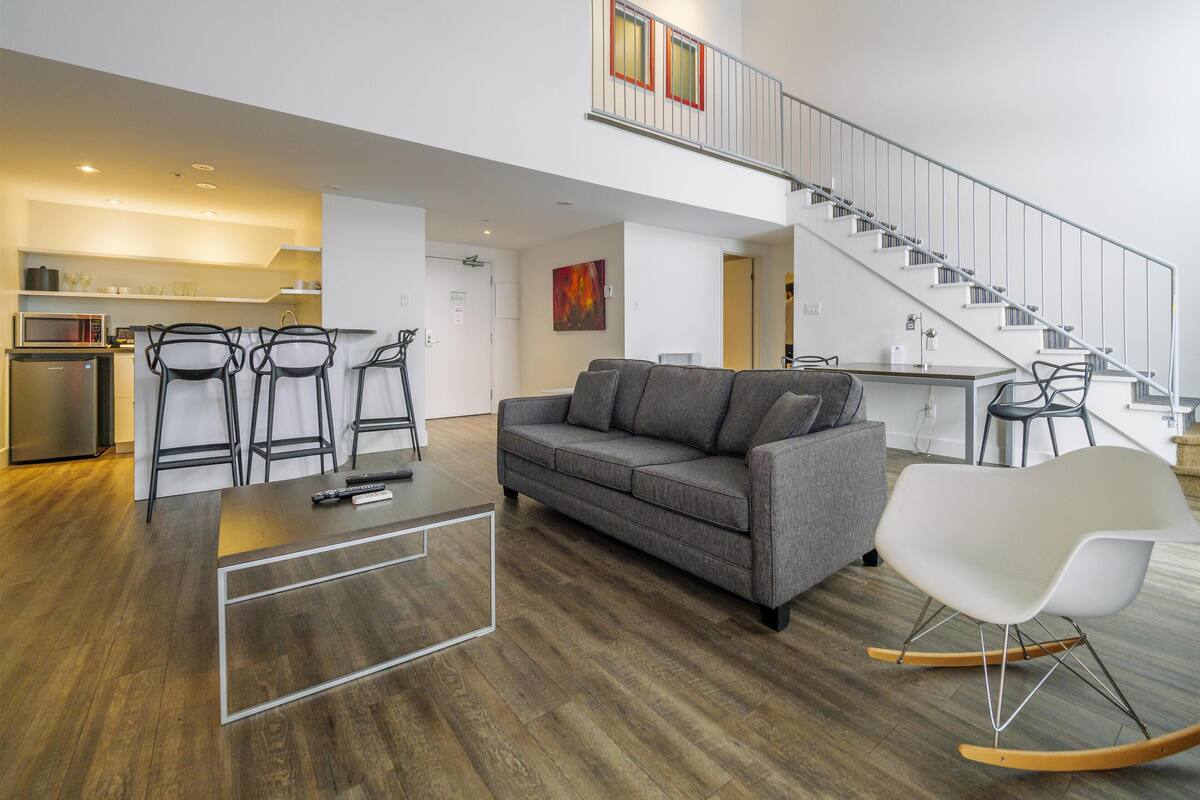 A modern living room with a grey sofa, a coffee table, and a rocking chair. There is a kitchen area with a microwave and a refrigerator. A staircase leads to an upper level.