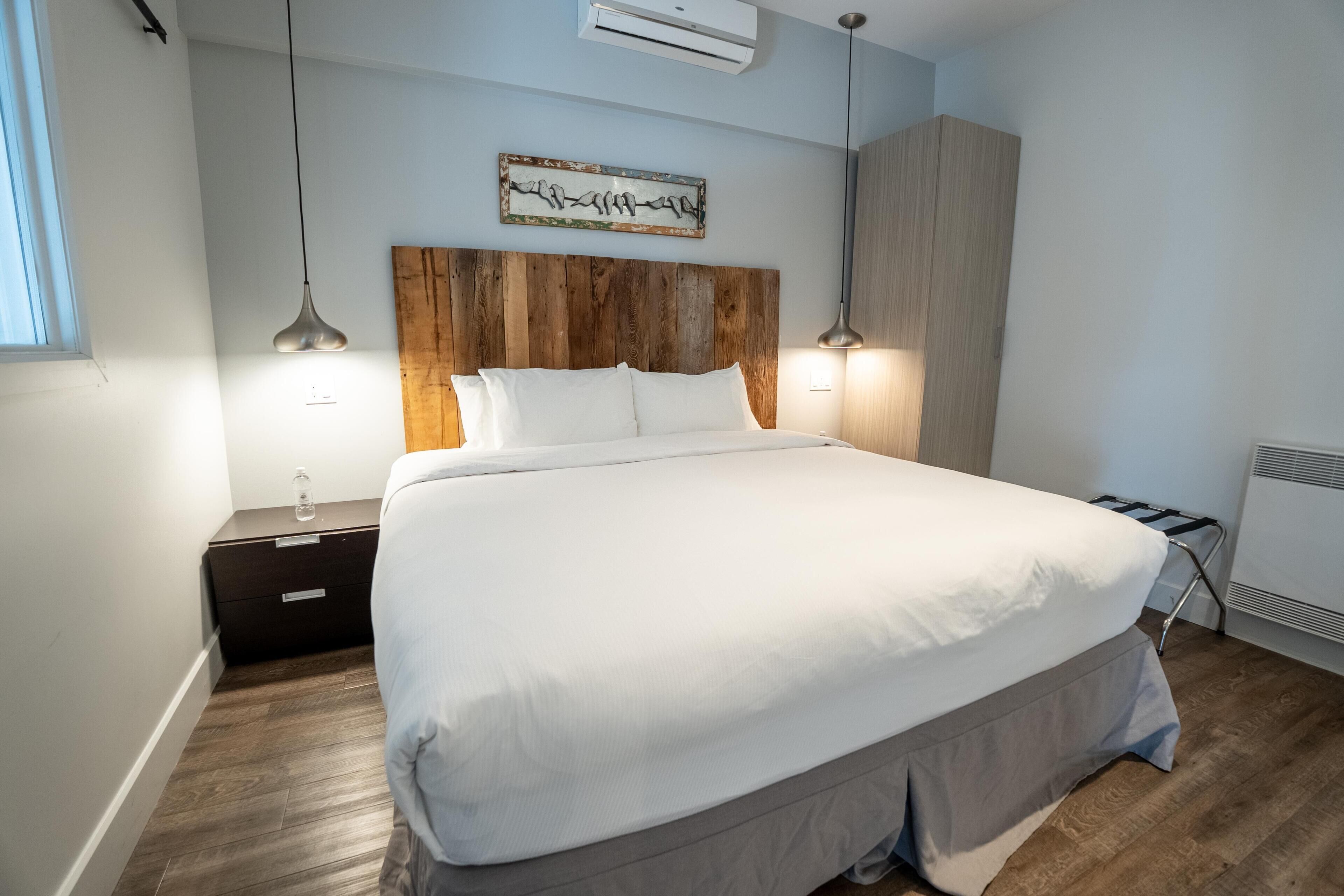 A modern bedroom with a wooden headboard, a large bed, two bedside lamps, and a wall-mounted air conditioner.