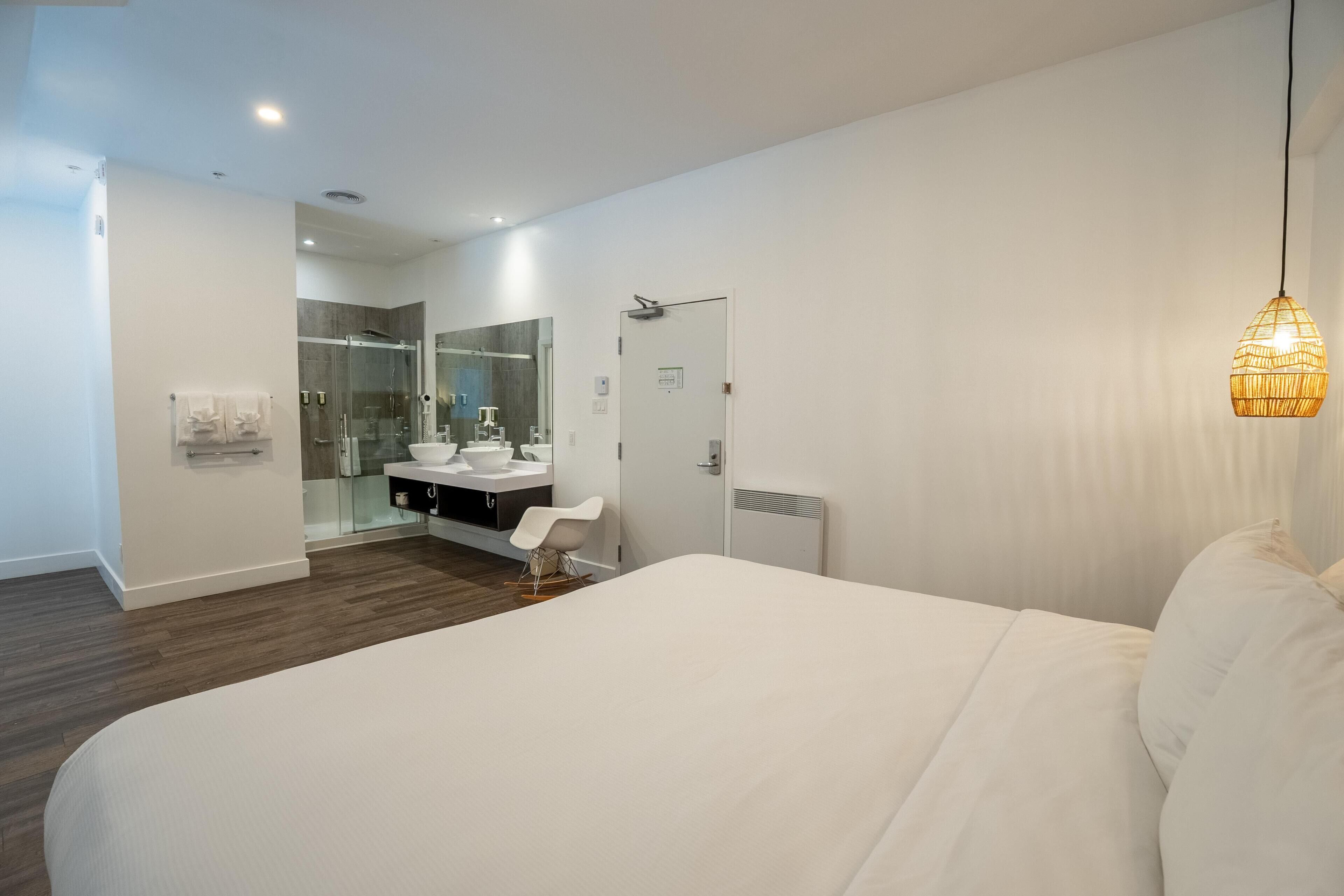 A modern bedroom with a large bed, a bedside table, a white chair, a bathroom with a glass shower enclosure, and a hanging light fixture.