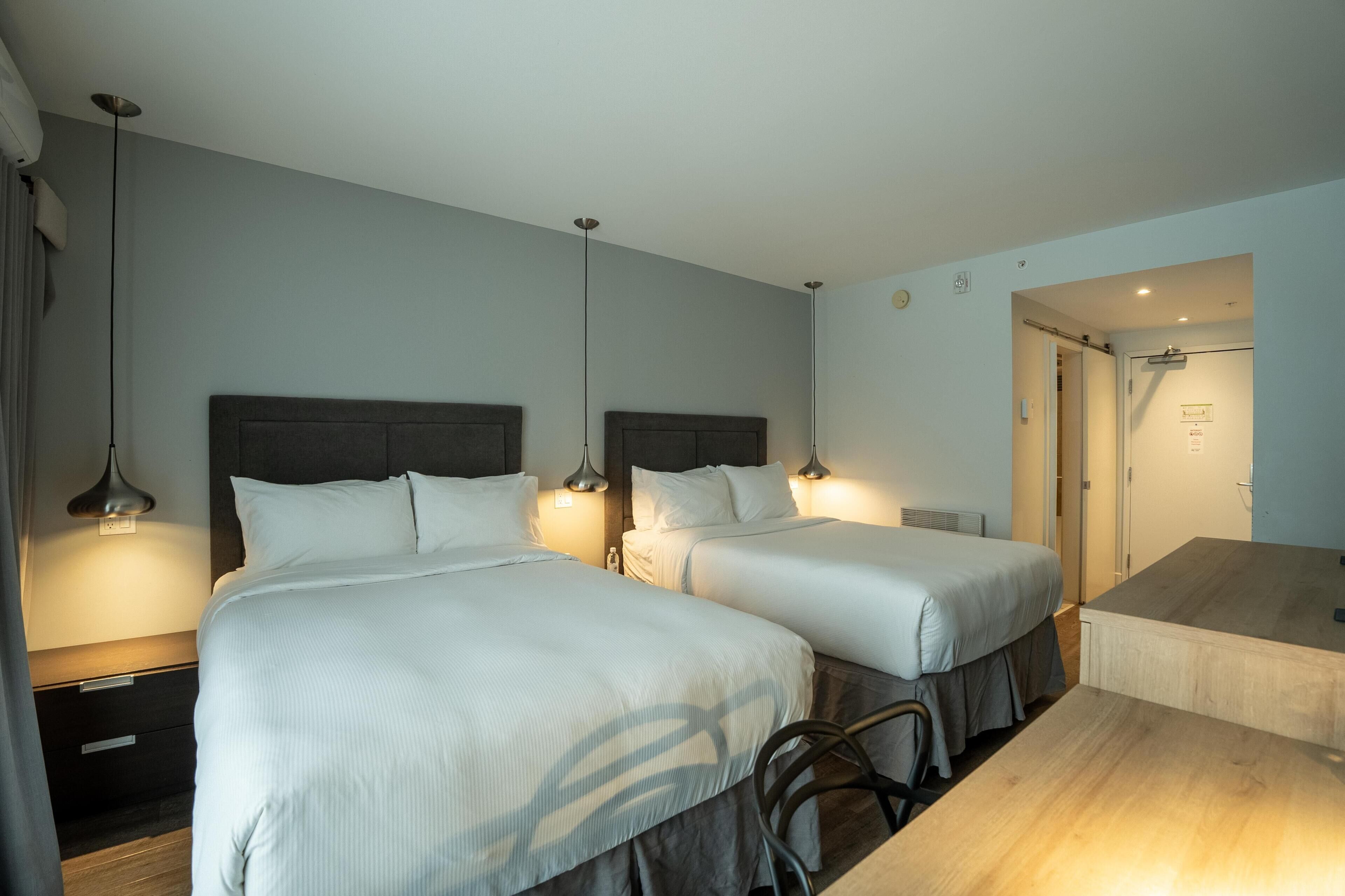 Habitación de hotel con dos camas, un escritorio y un baño visible a través de una puerta abierta.