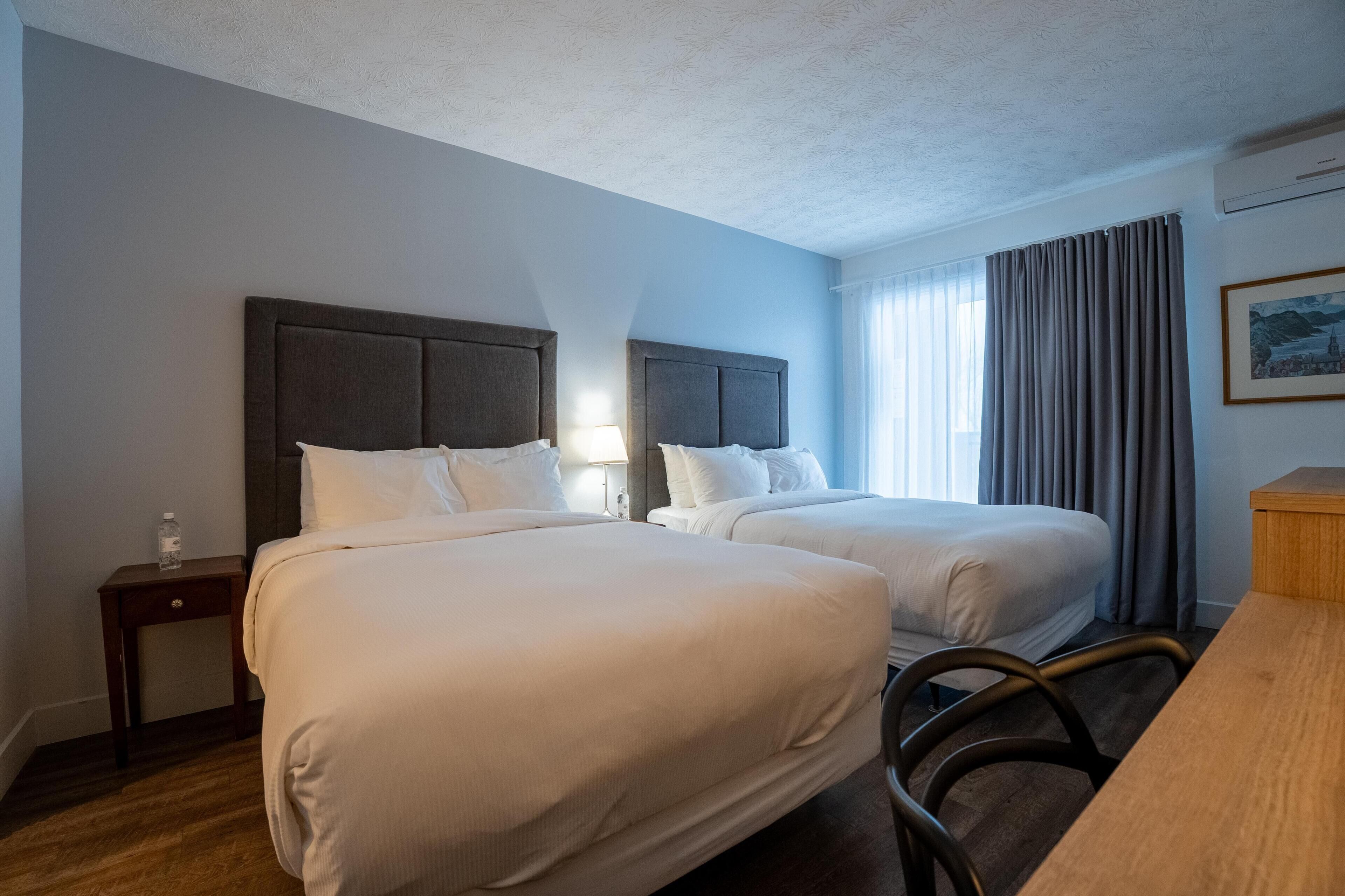 A hotel room with two beds, a wooden desk, a nightstand, and a window with curtains.