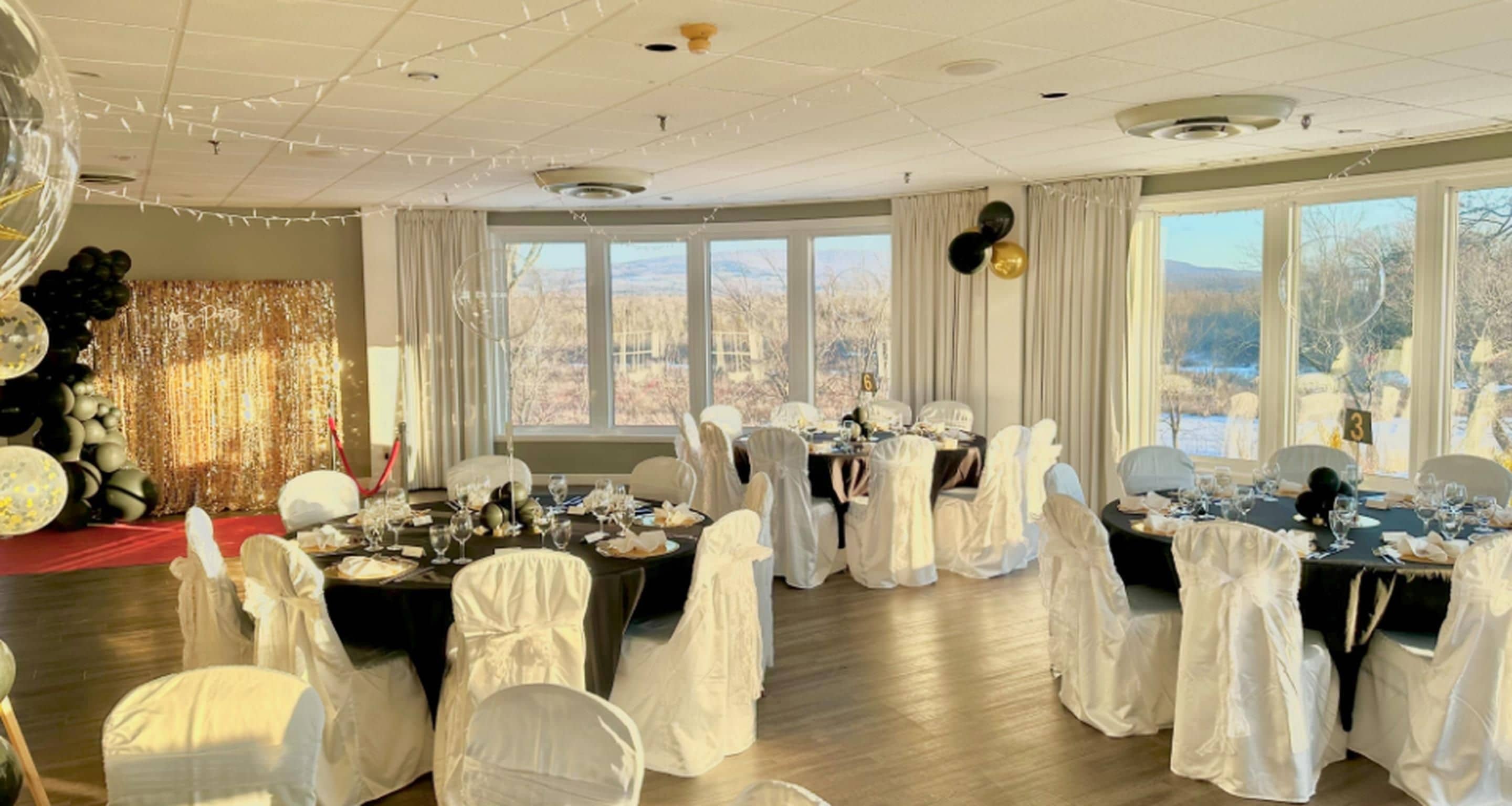 A banquet hall with tables set for a formal event, large windows offering a view of the outdoors, and a ceiling with decorative lighting.