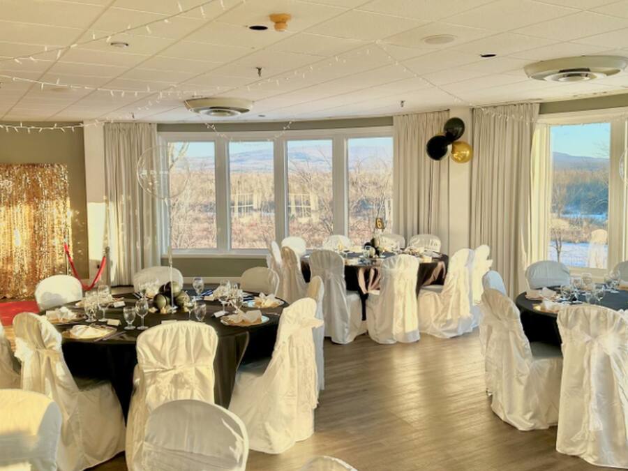 A banquet hall with tables set for a formal event, large windows offering a view of the outdoors, and a ceiling with decorative lighting.