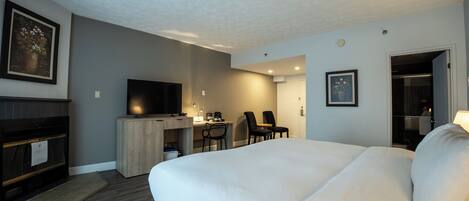 Quarto de hotel com uma cama grande, uma escrivaninha com computador, uma televisão e duas cadeiras.