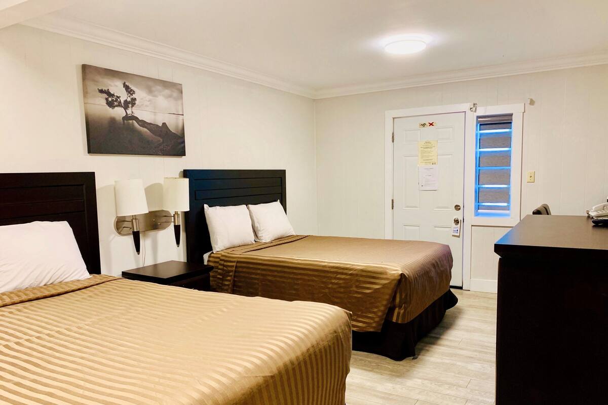 A hotel room with two beds, a desk, a lamp, a painting on the wall, and a door with a window.